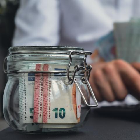 Money in a jar and a person writing on a computer holding more money