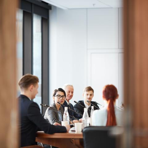 Men and women sitting at a conference room table with mics, discussing with each other.